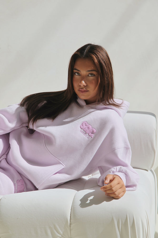Woman wearing the hoodie, seated on a white boucle chair. Cosy Club 2.0 Adults Hoodie in Lilac by Skylaroseco