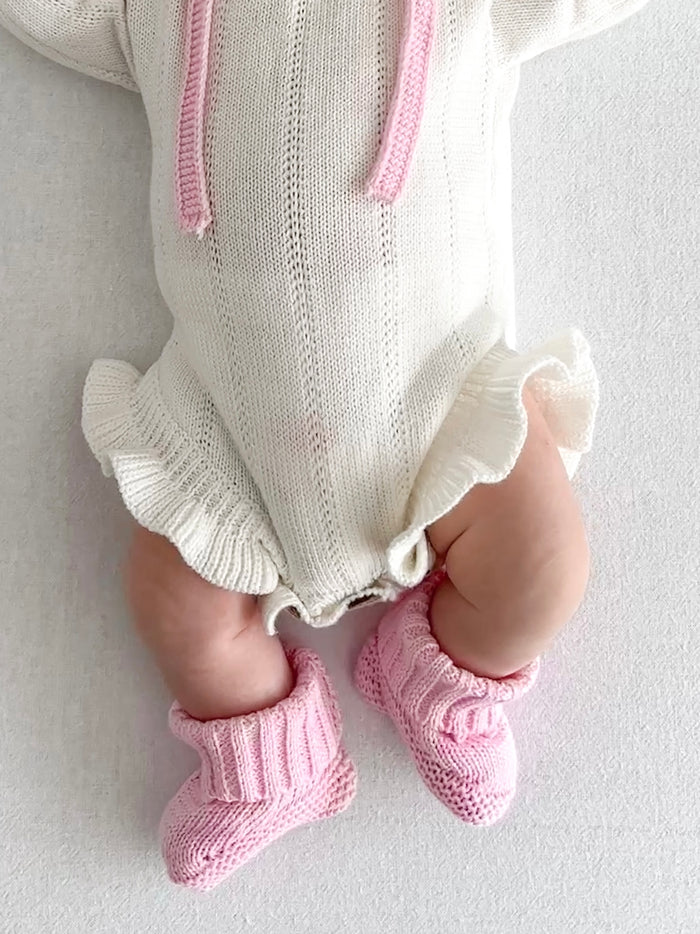 Close-up of a baby wearing the booties with a white knit romper. Knitted Booties in Pink by Skylaroseco