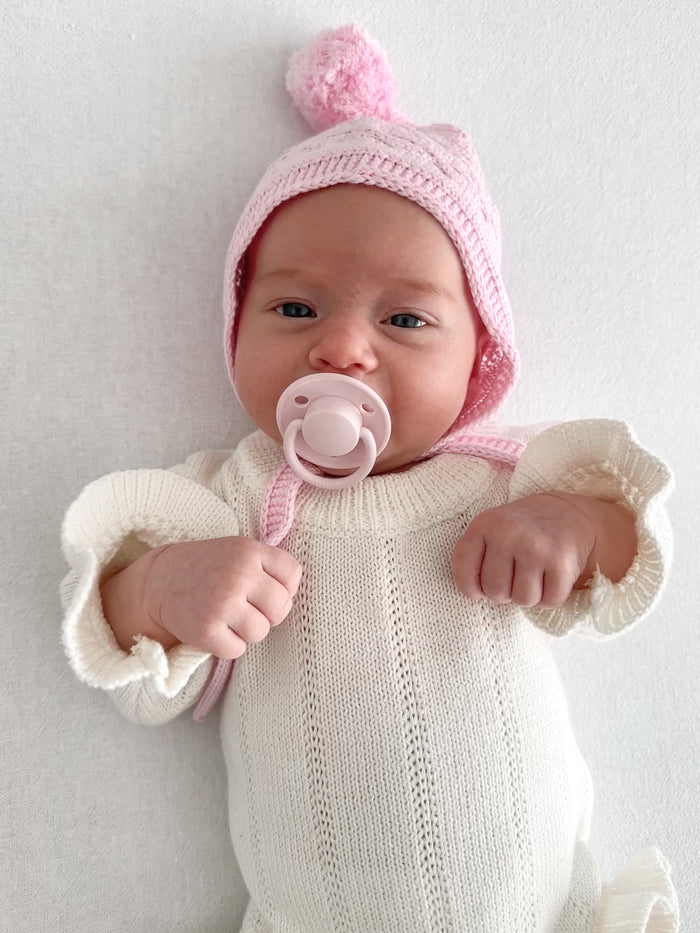Baby wearing the bonnet with a dummy and a white knit romper. Knitted Pixie Bonnet in Pink by Skylaroseco