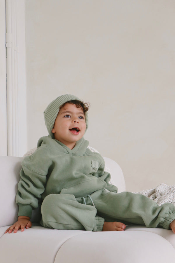 Smiling toddler sitting on a boucle chair in the sage lounge set and beanie. Cosy Club 2.0 Kids Lounge Set in Sage by Skylaroseco