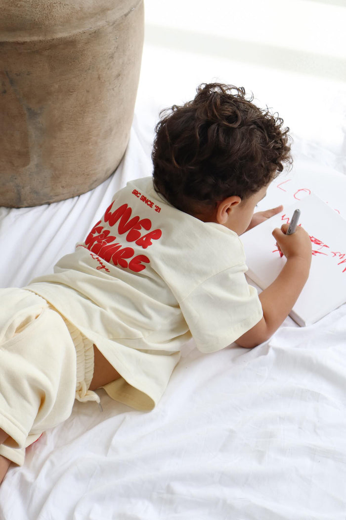 Child lying on white bedding drawing, wearing the cream tee with red print. Love & Leisure Kids Oversized Tee in Cherry Cream by Skylaroseco