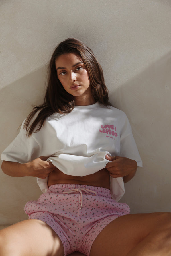 Woman seated in the white oversized tee with pink print and pink floral shorts. Love & Leisure Adults Oversized Tee in Bon Bon by Skylaroseco